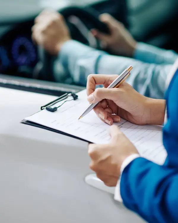 A professional dressed in business attire fills out paperwork on a clipboard while seated inside a vehicle, suggesting documentation review or registration processing during a vehicle inspection.