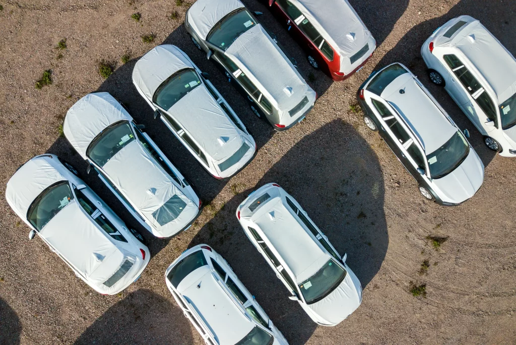 An aerial view of a fleet of predominantly white SUVs staged in organized rows on a gravel lot, likely awaiting bulk registration, titling, or usement processing.