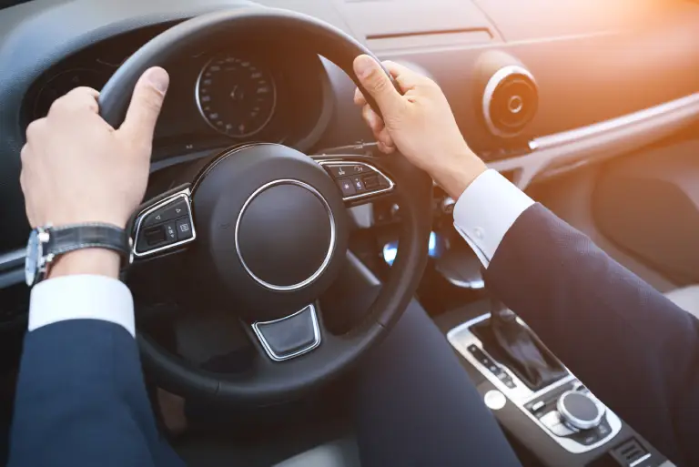 Business professional driving a modern vehicle, hands on steering wheel, interior dashboard and center console visible with sunlight entering through the windshield.