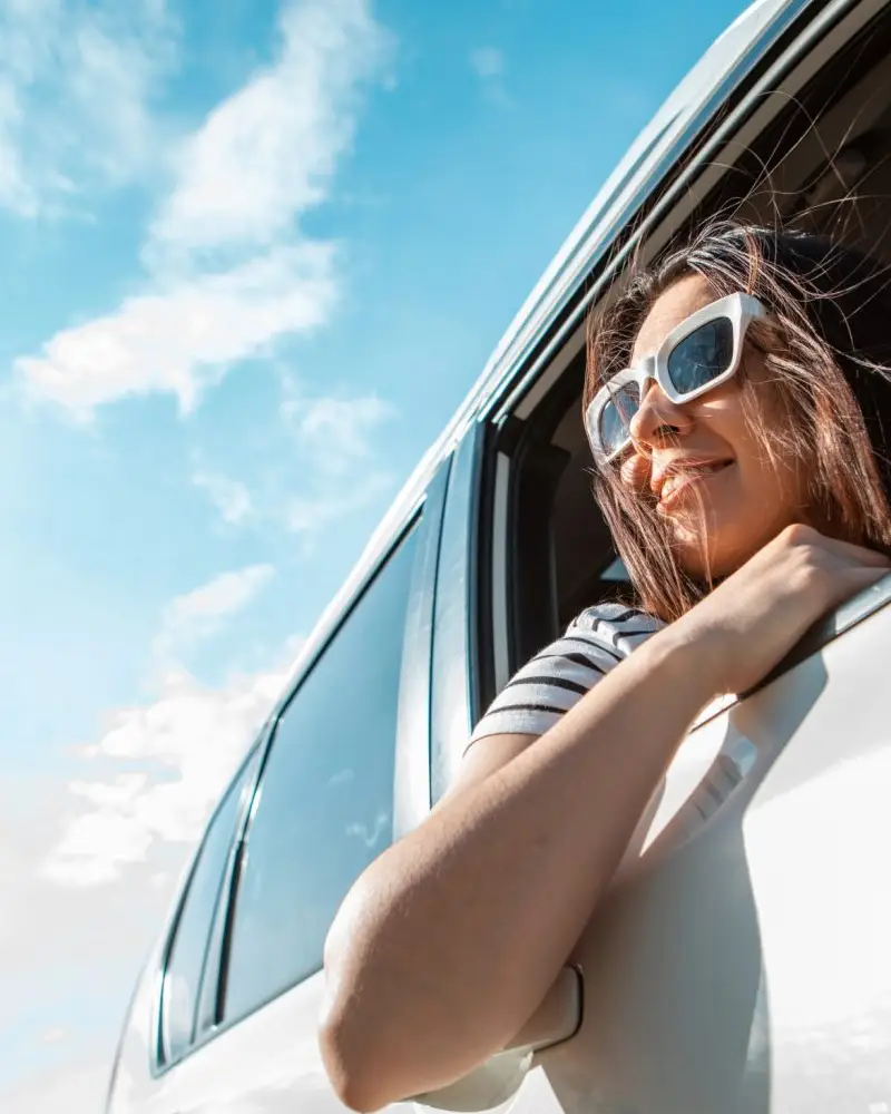 A woman wearing sunglasses smiles out the window of a vehicle on a sunny day, enjoying the freedom of the open road during a road trip.