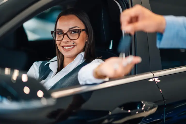 A smiling woman sitting in the driver’s seat of a car receives keys through the window, symbolizing LLC MT services and vehicle registration assistance.