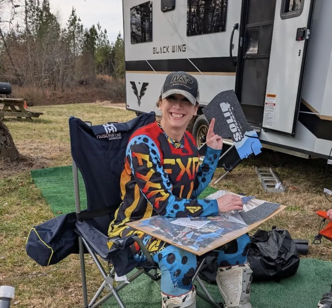 Crystalmarie smiling and giving a thumbs up while sitting in a camp chair in full motorcycle gear next to her Black Wing toy hauler