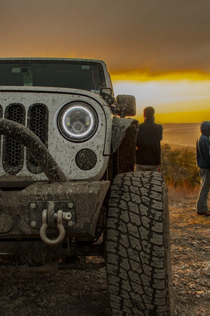 Muddy Jeep Wrangler parked on dirt road at sunset with people standing nearby discussing off-road vehicle registration