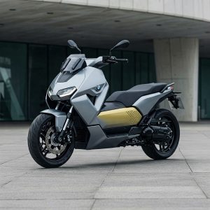 A modern gray electric scooter-style motorcycle parked on a paved plaza in front of a contemporary building with large glass windows.