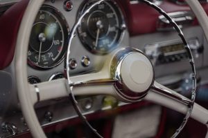 A close-up view of a vintage car interior featuring a classic steering wheel with a chrome horn ring and analog dashboard gauges set against rich red upholstery.