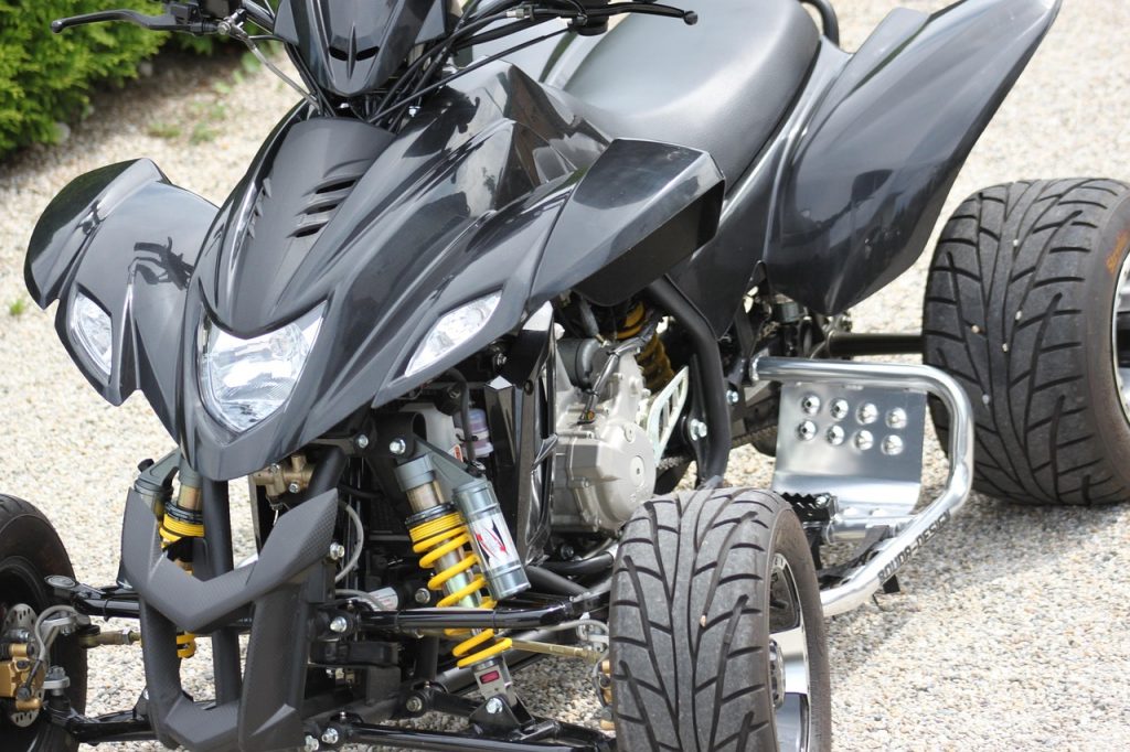 Modified black quad ATV with yellow suspension, LED lights, and street tires parked on gravel