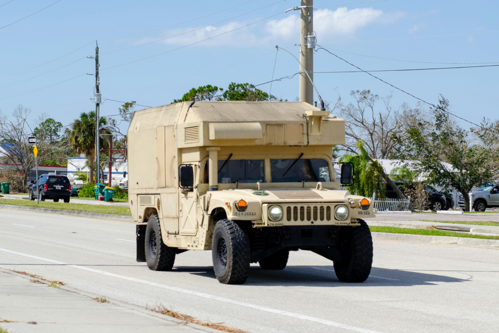 Tan military Humvee truck with custom camper cab conversion driving on suburban street during daytime.