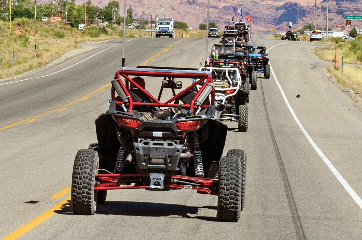 Sideway1_DSC_2865 A group of customized side-by-side UTVs drives in a line down a two-lane desert highway, surrounded by rocky hills and open landscape.