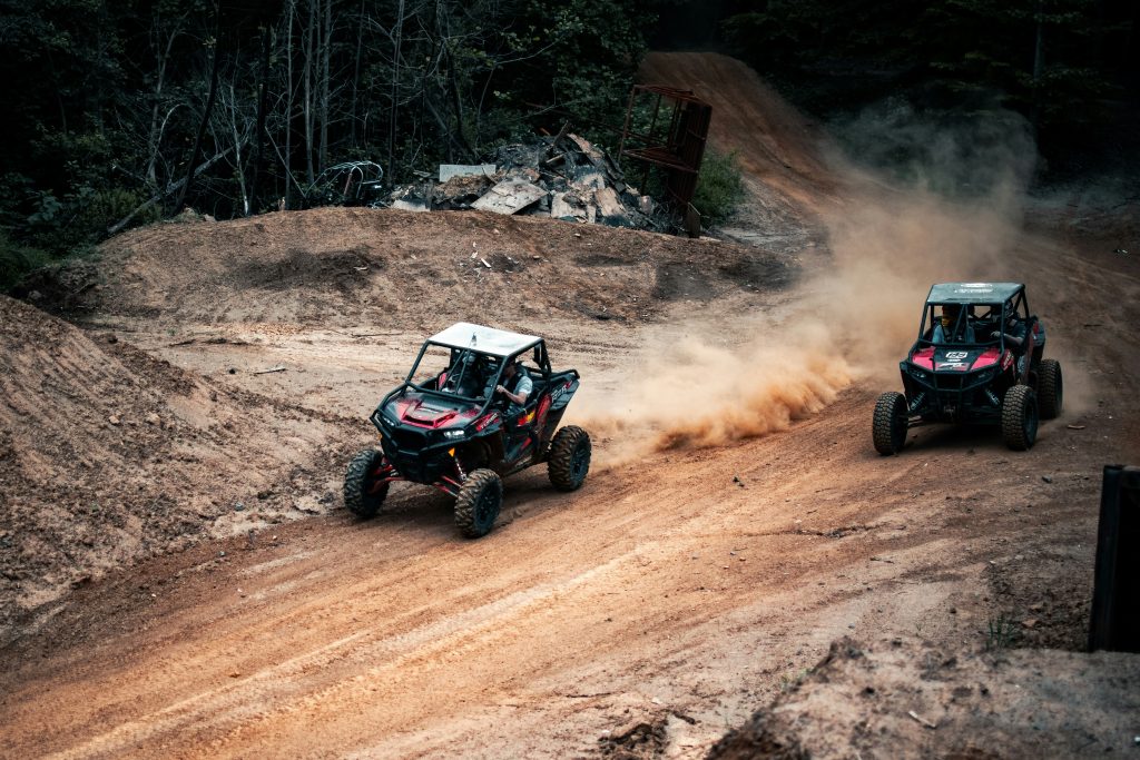 Two off-road UTVs racing through dusty dirt terrain with forest backdrop, demonstrating recreational vehicle riding on trail.