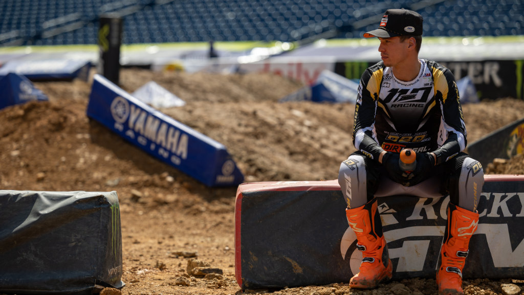 A Dirt Legal DBDRacing WMR KTM rider in black, gold, and white gear with orange boots and a hat sits on a track barrier. He looks thoughtfully toward the distance at a Supercross stadium.