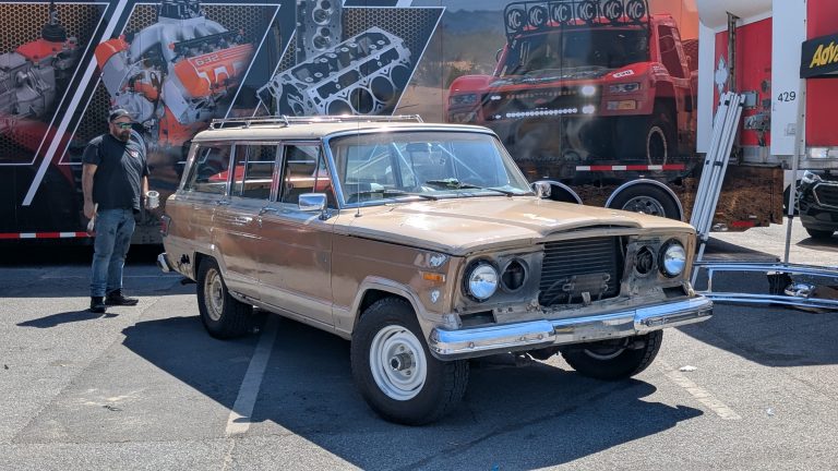 Classic Jeep Wagoneer parked at an outdoor automotive event with custom engine display trailer and performance truck graphics in the background.