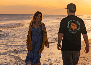 A man and woman walking along the shoreline at sunset, with gentle waves reflecting the golden light and the ocean stretching out behind them.
