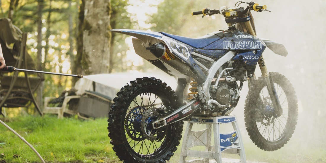 Spring-Clean-Dirt-Bike A blue Yamaha dirt bike is being pressure washed on a stand outdoors, surrounded by trees and warm sunlight, getting cleaned up and ready for spring riding season.