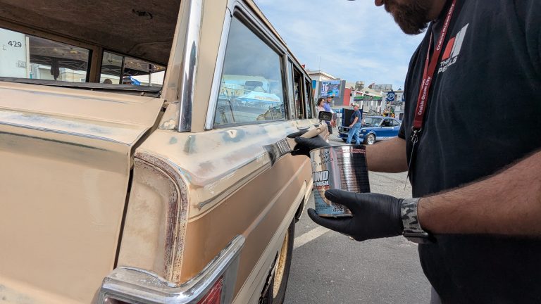 Person wearing gloves applying polish to the side of a classic Jeep Wagoneer during an outdoor car show event.