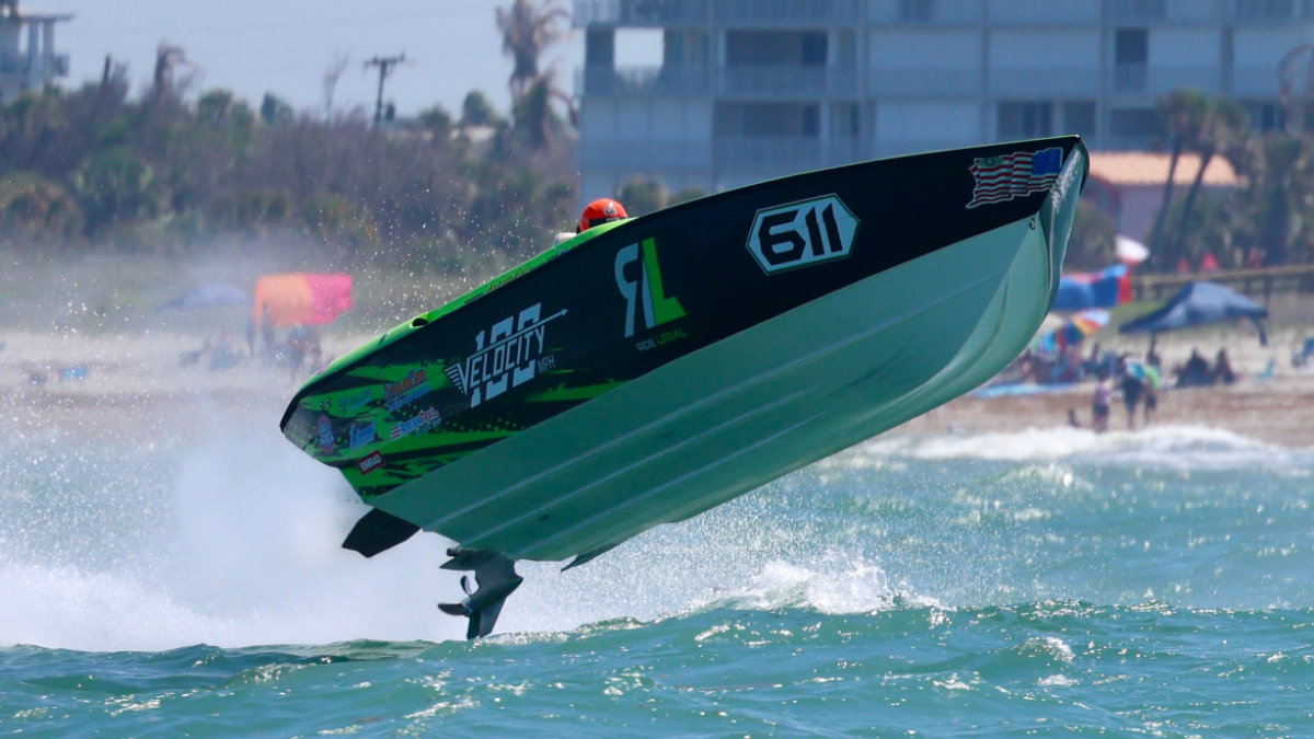 Green speedboat launches out of the water during a seaside race, with beachgoers and buildings in the background.