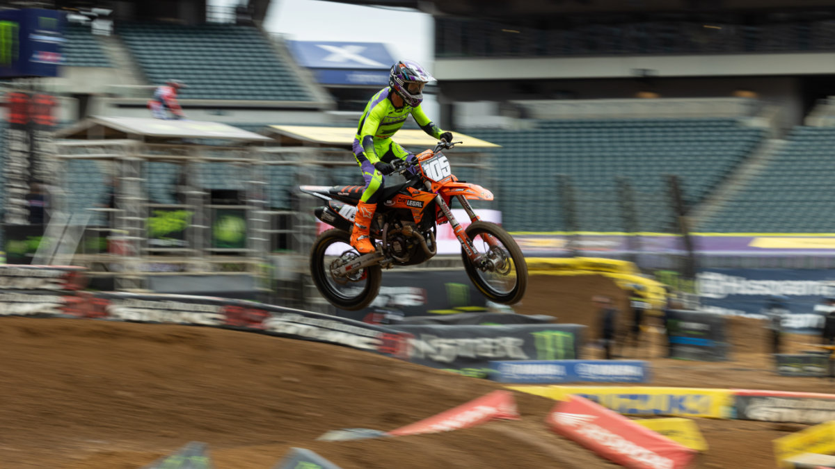 A Dirt Legal sponsored professional motocross rider in bright neon green gear executes a big jump during a race in a stadium.