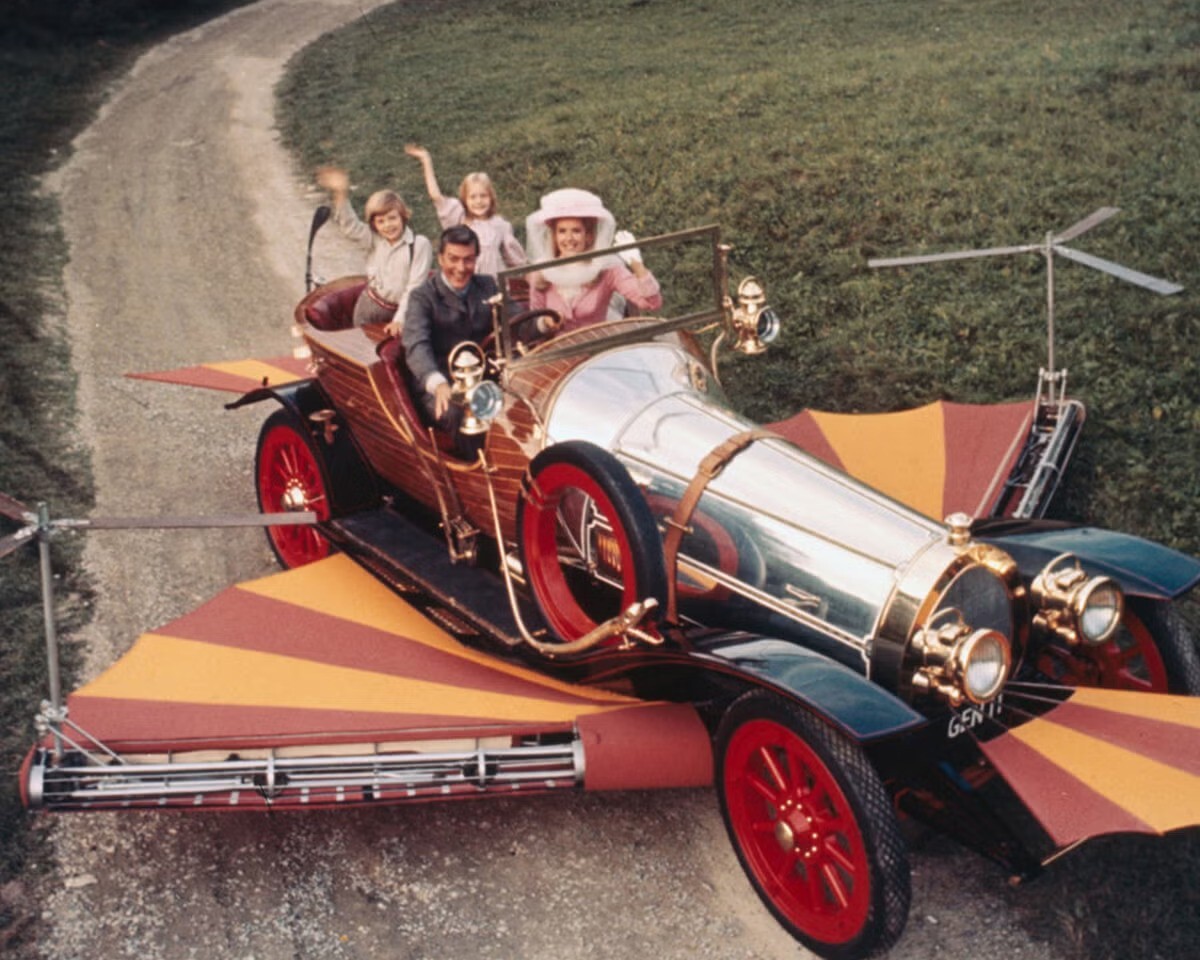 A family of four rides in the whimsical flying car from Chitty Chitty Bang Bang, complete with striped airplane-style wings and a rear propeller, traveling along a country road as the children wave excitedly during their magical adventure.