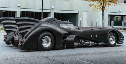 A sleek, matte-black Batmobile-inspired car with dramatic fins, oversized rear wheel covers, and exposed mechanical components is parked on a city street in front of a modern building, showcasing an aggressive, futuristic design.