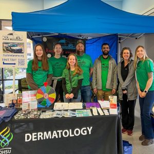 Group of people in green shirts standing behind a dermatology information booth under a blue canopy at a community event.