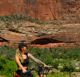 A woman with long curly hair and visible arm tattoos standing beside a bicycle in a desert landscape, with towering red rock cliffs and greenery in the background.