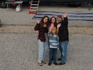 A family of four standing together on a gravel campsite, smiling and waving at the camera with an RV parked behind them.