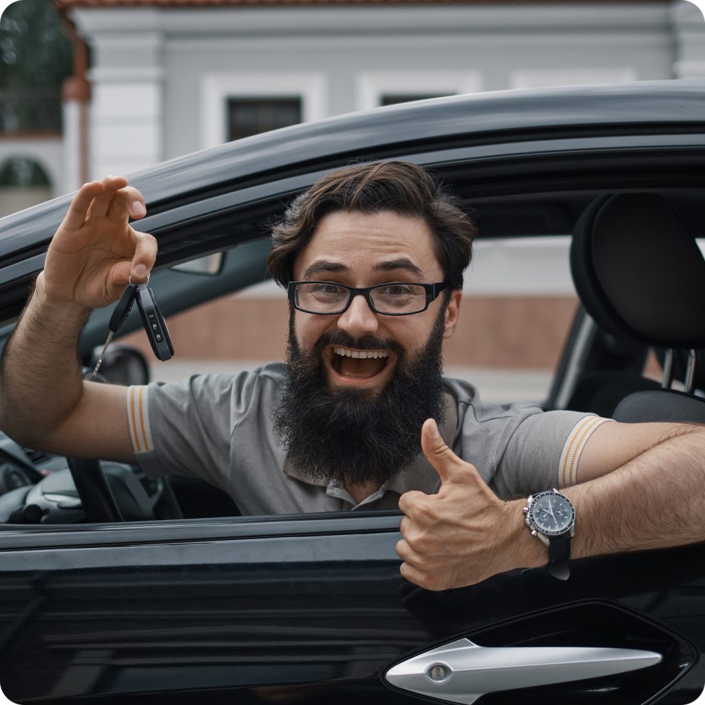A smiling man sits in his car holding up the keys and giving a thumbs-up, representing someone who has successfully formed a Montana LLC and registered their vehicle with ease.