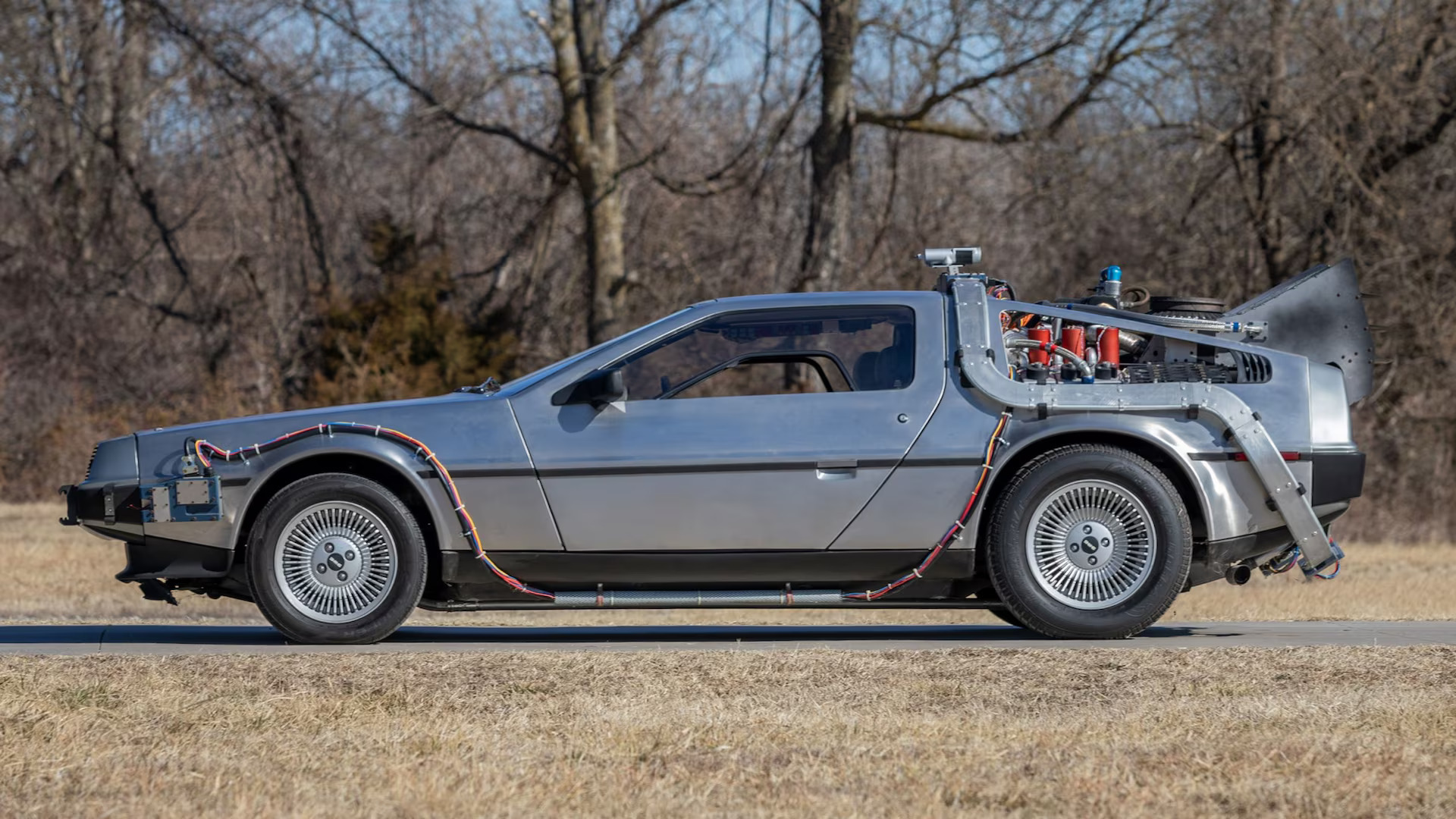 A silver DeLorean DMC-12 with exposed wiring and a large rear-mounted time machine apparatus is parked on a roadside, showcasing its iconic stainless steel body and futuristic modifications inspired by Back to the Future.