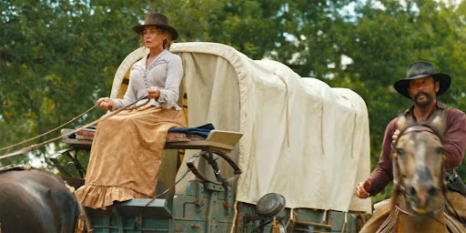 A woman in pioneer-era clothing drives a covered wagon pulled by horses while a man on horseback rides alongside her, set against a wooded background that evokes a journey through the Old West.