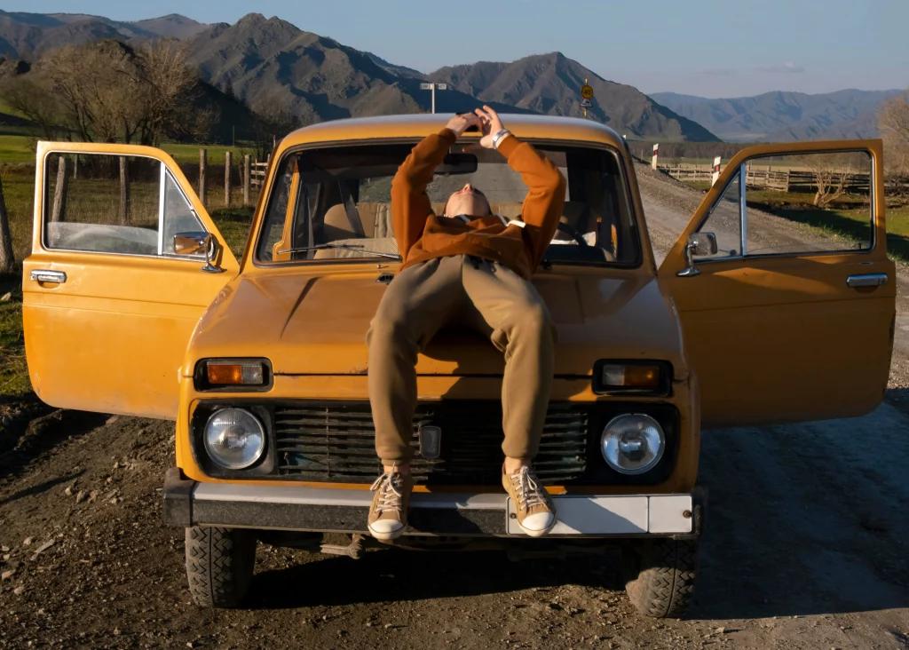 Person relaxing on the hood of a vintage orange off-road vehicle parked on a rural dirt road with mountain views in the background. The image highlights the connection between slowing down, mental well-being, and enjoying the freedom that comes with the open road.