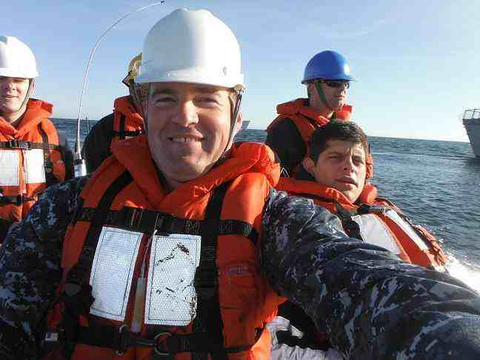 A group of U.S. Navy personnel wearing camouflage uniforms, life jackets, and safety helmets are riding in a small boat on the open water, smiling and taking a selfie during what appears to be a training or transport mission.