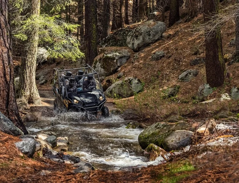 Black Kawasaki UTV splashing through creek water in forested mountain terrain with rocks and tall trees