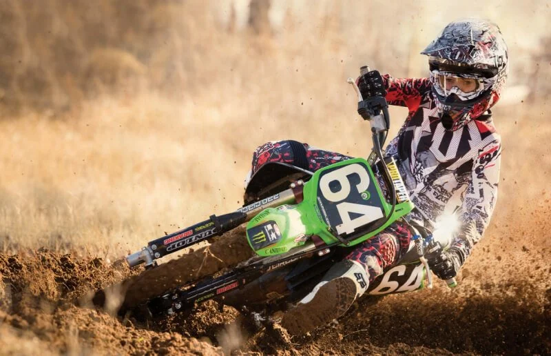 Young rider on green dirt bike leaning into turn on dusty track, kicking up dirt clouds during off-road riding.