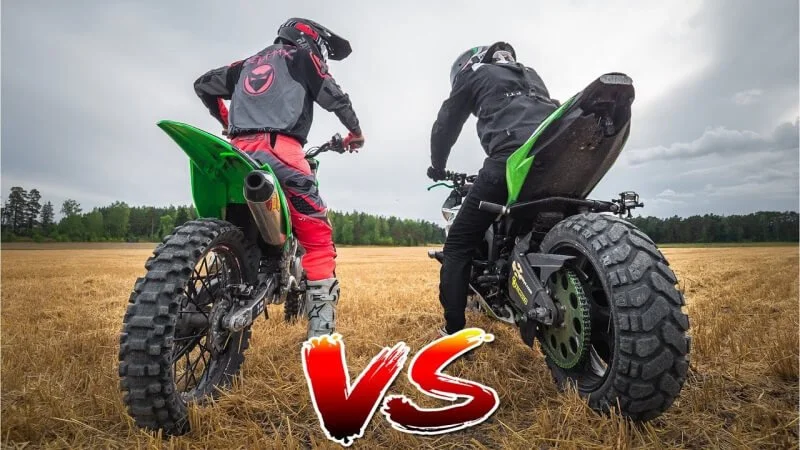 Two dirt bike riders standing on their motorcycles in open field facing off in competitive stunt comparison