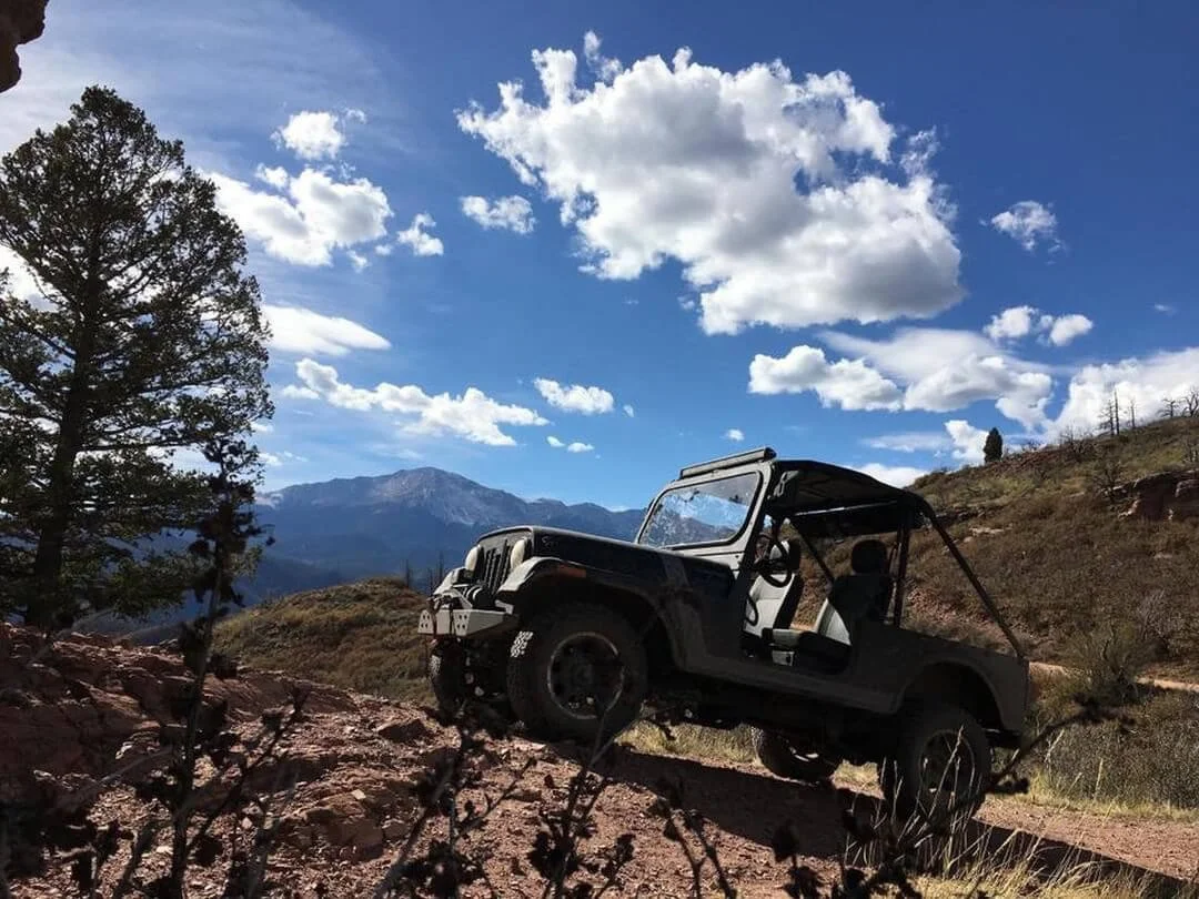Jeep parked on mountain hillside with door open, scenic landscape and blue sky background, off-road vehicle adventure
