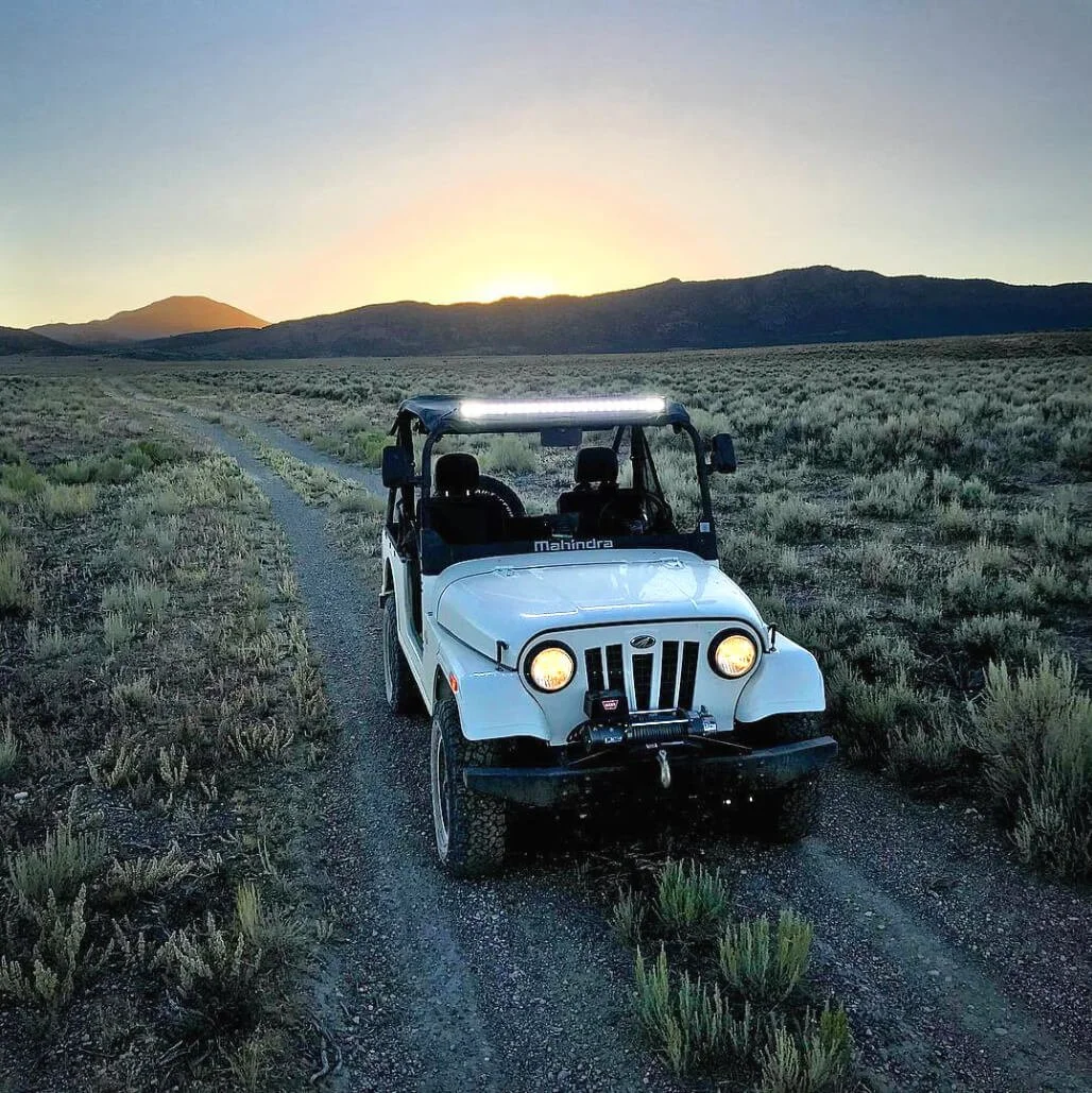White Mahindra Roxor off-road vehicle with headlights on traversing desert terrain at dusk with mountains
