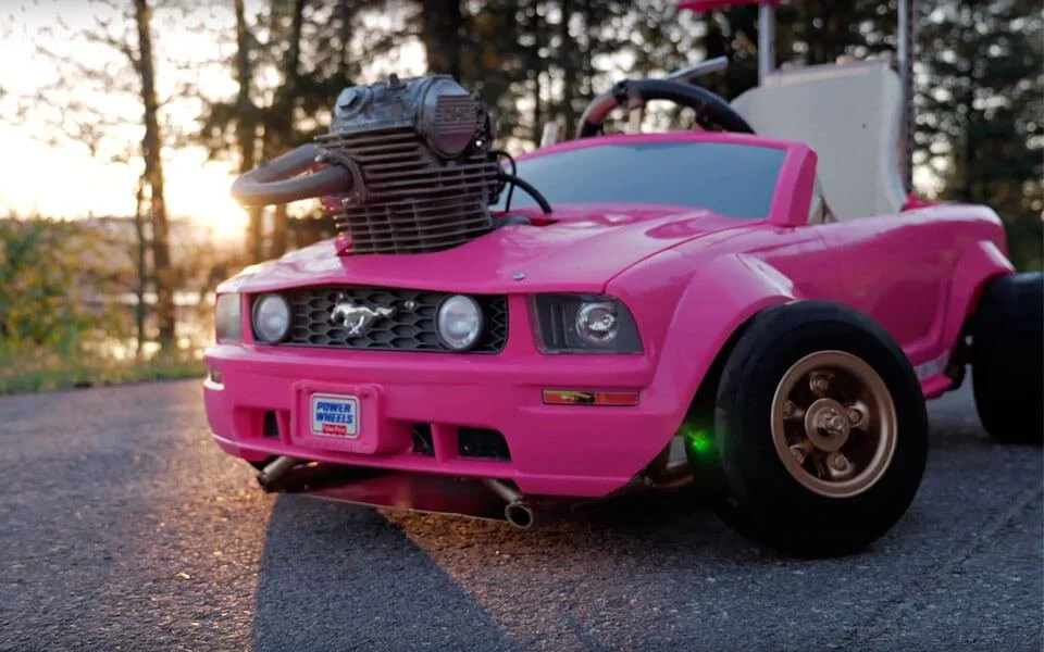 Pink Mustang children's ride-on car with motorcycle engine swap conversion parked outdoors at sunset