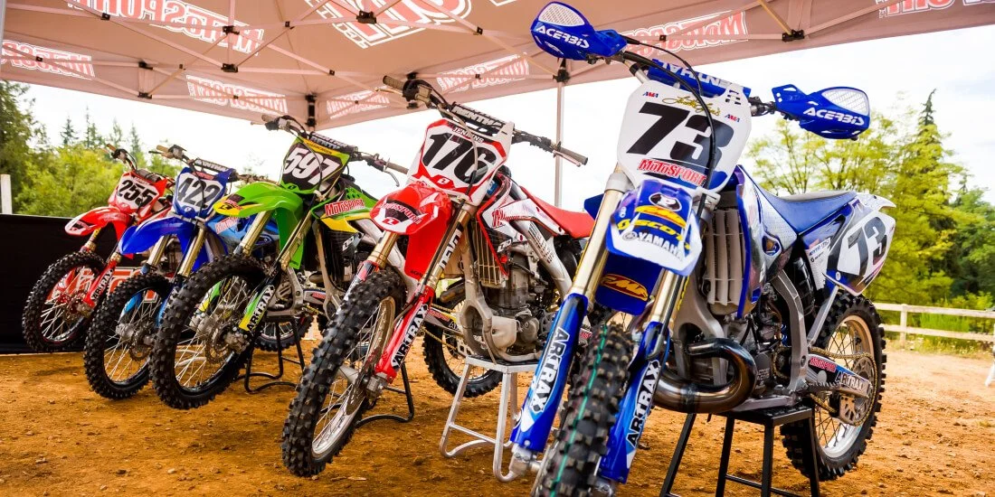 Professional dirt bikes lined up under tent display, featuring red, blue, and green beginner-friendly motocross models
