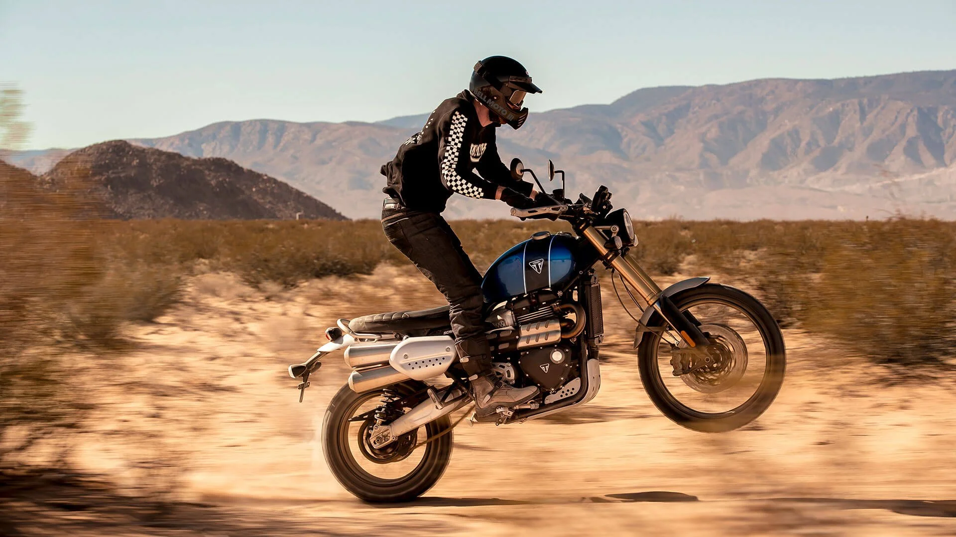 Rider performing wheelie on blue and black Triumph Scrambler 1200 motorcycle in desert landscape