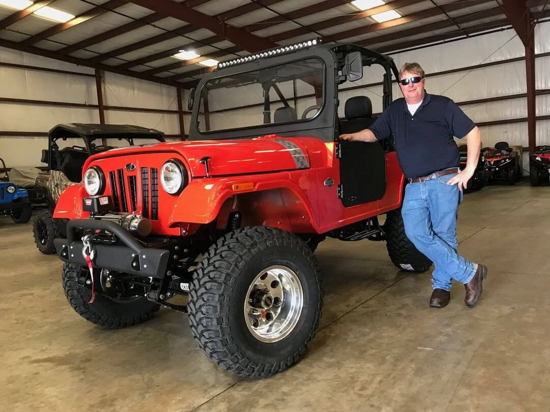 Red lifted Jeep-style Roxor vehicle with oversized tires displayed in warehouse garage setting with owner