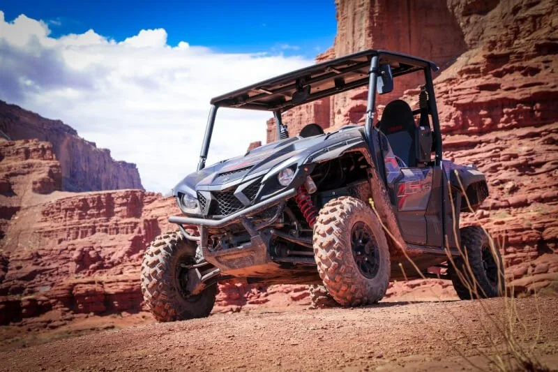 Red Yamaha UTV parked on desert sand with red rock canyon formations and blue sky in background