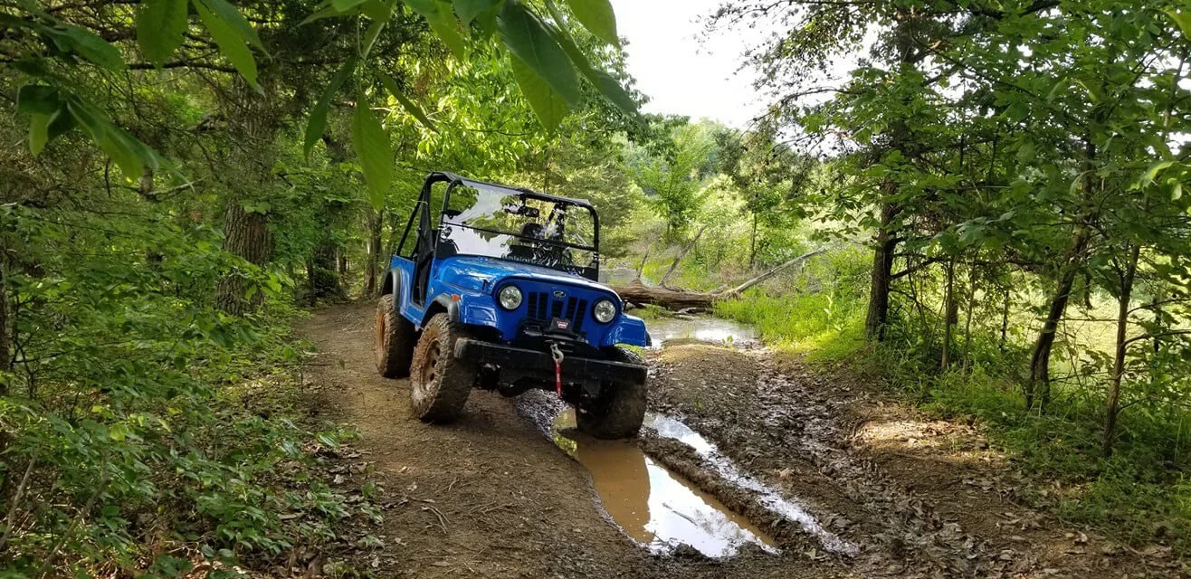 Blue Roxor off-road vehicle navigating muddy forest trail through woodland terrain during extreme testing.