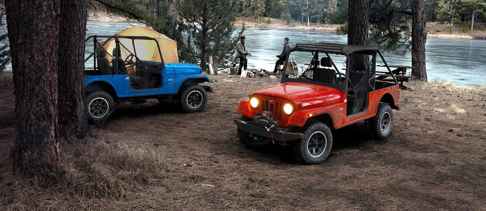 Two colorful Mahindra Roxor vehicles parked at a riverside camping site with trees and water visible in background.
