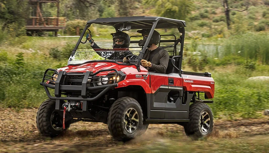 Red Kawasaki Mule utility vehicle driven by two people on forest trail with fire safety recall concerns.