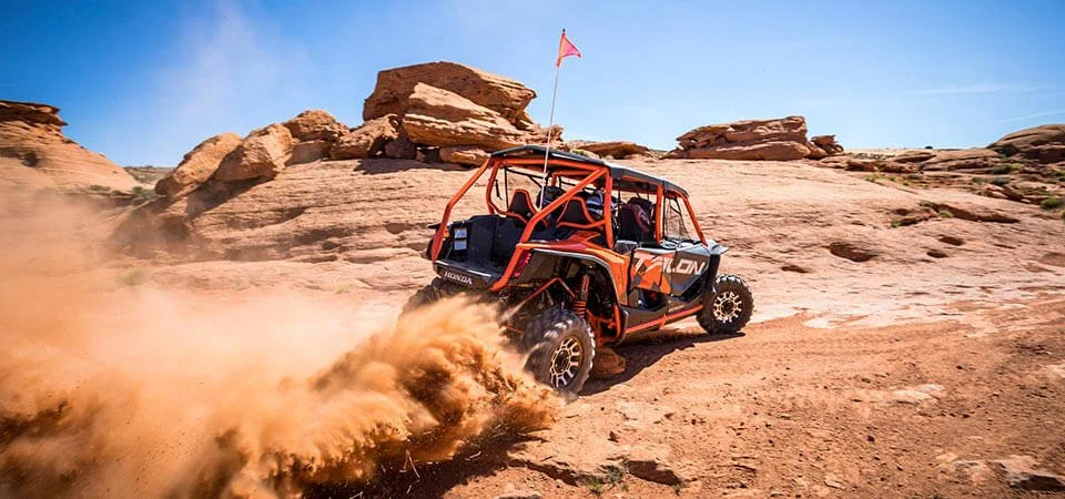Red four-seater Honda Talon UTV kicks up dust while driving through rocky desert terrain with flag mounted on roll cage