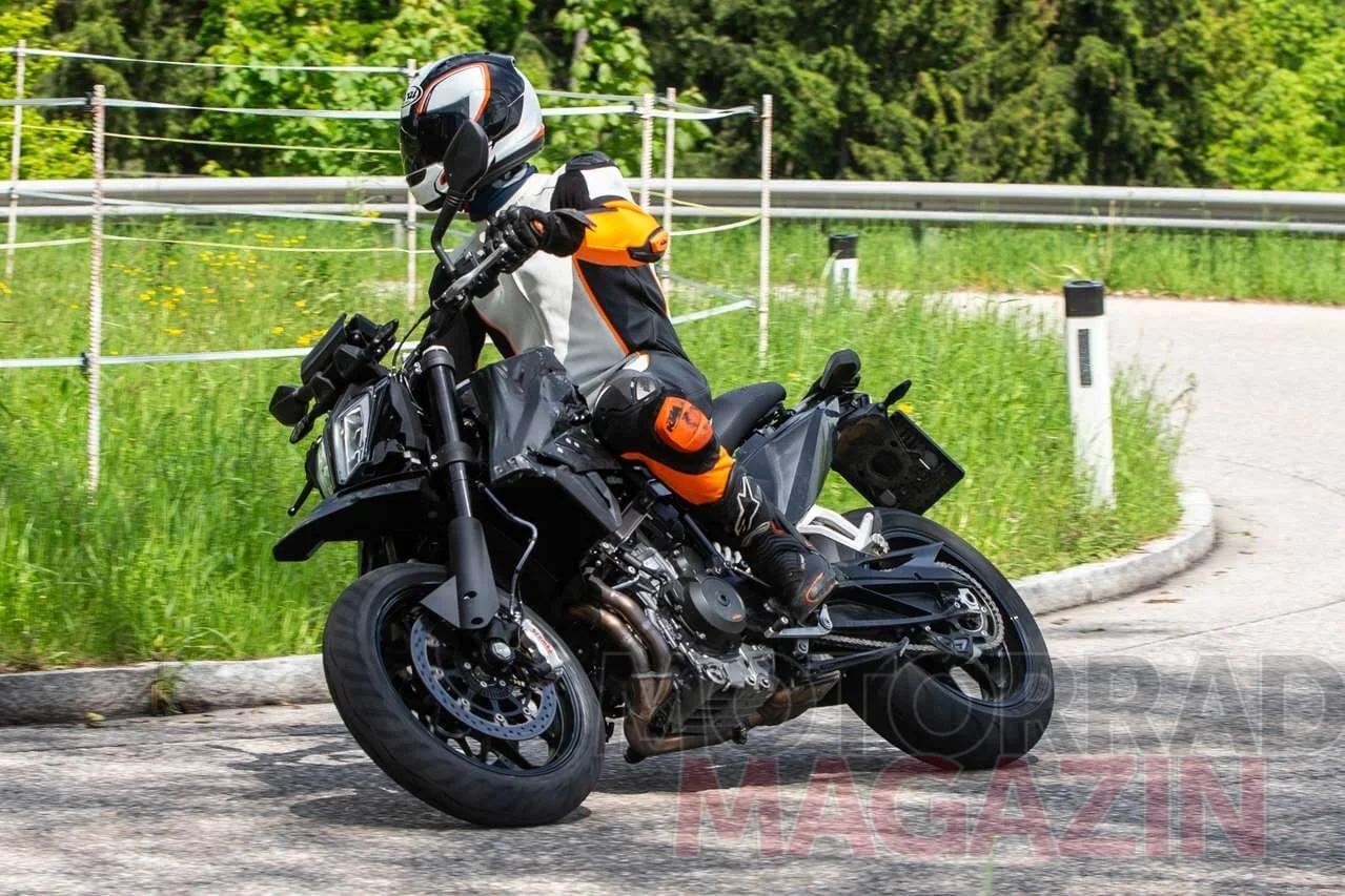 Rider on orange and black KTM 790 SMC supermoto motorcycle performing wheelie on paved track with green grass background