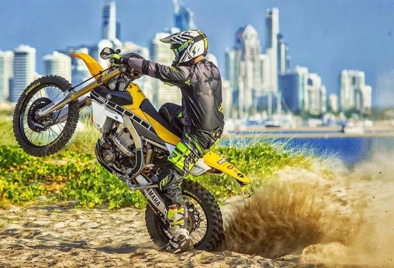 Yellow dirt bike rider performing wheelie on sandy beach with city skyline backdrop