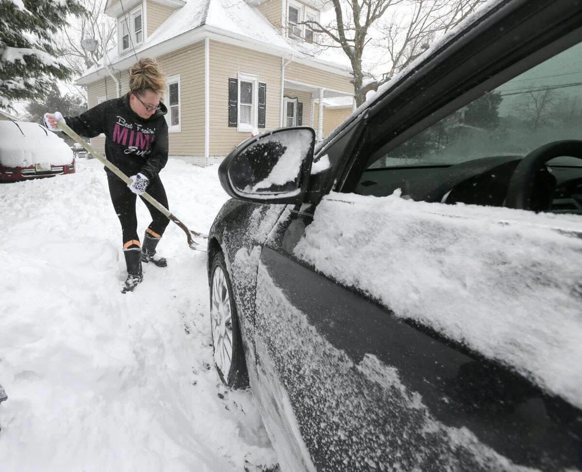 Woman shoveling snow from car in driveway during winter weather for vehicle maintenance safety