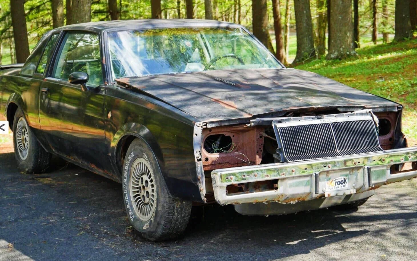 Damaged car with missing front end and exposed engine displayed for parts only in wooded driveway
