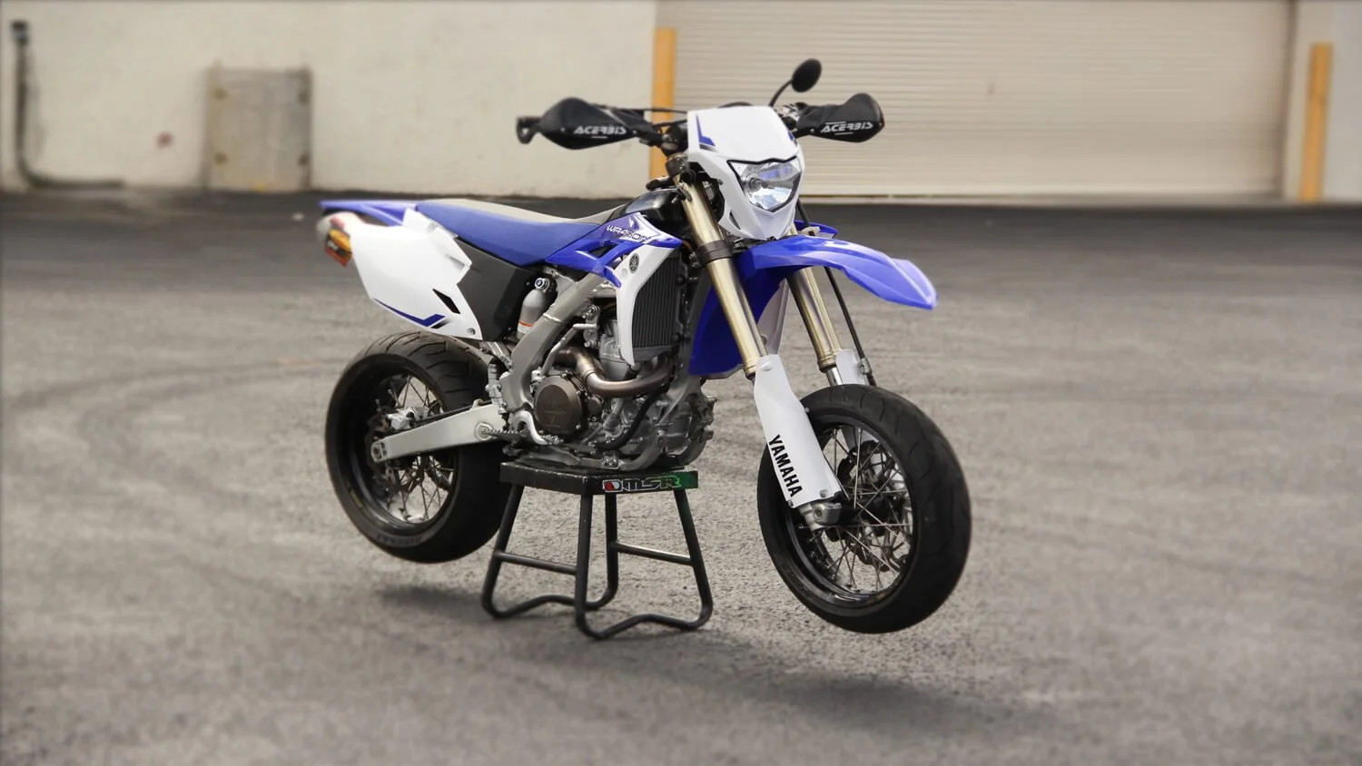 Blue and white street legal dirt bike displayed on maintenance stand in parking garage