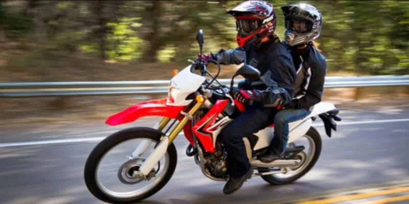 Two riders on a red dirt bike modified for street legal use riding on California highway with trees blurred in background
